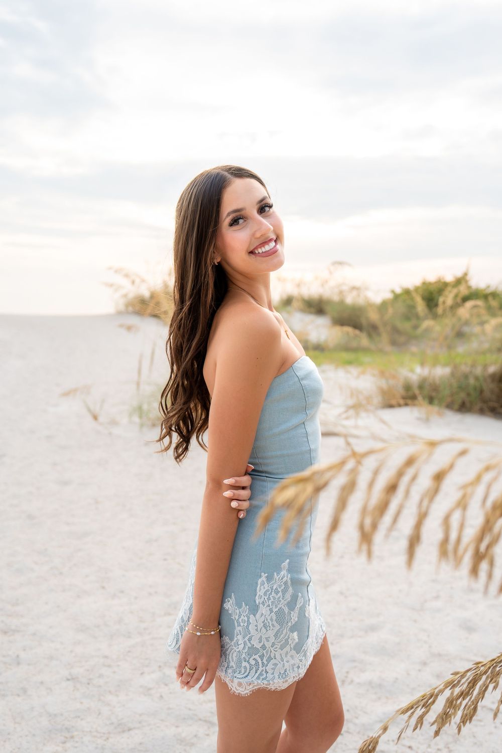 Smiling brunette woman in a light blue strapless lace dress standing on a sandy beach at sunset.