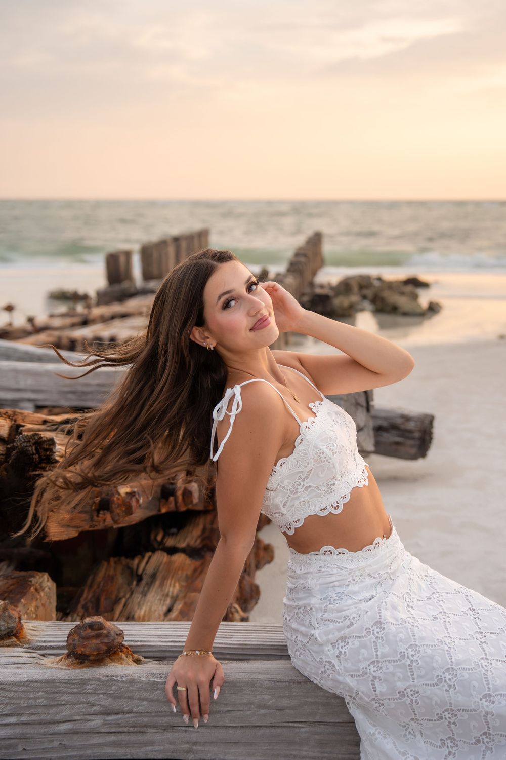 Woman in white lace beach outfit leaning on driftwood with wind-blown hair at sunset.