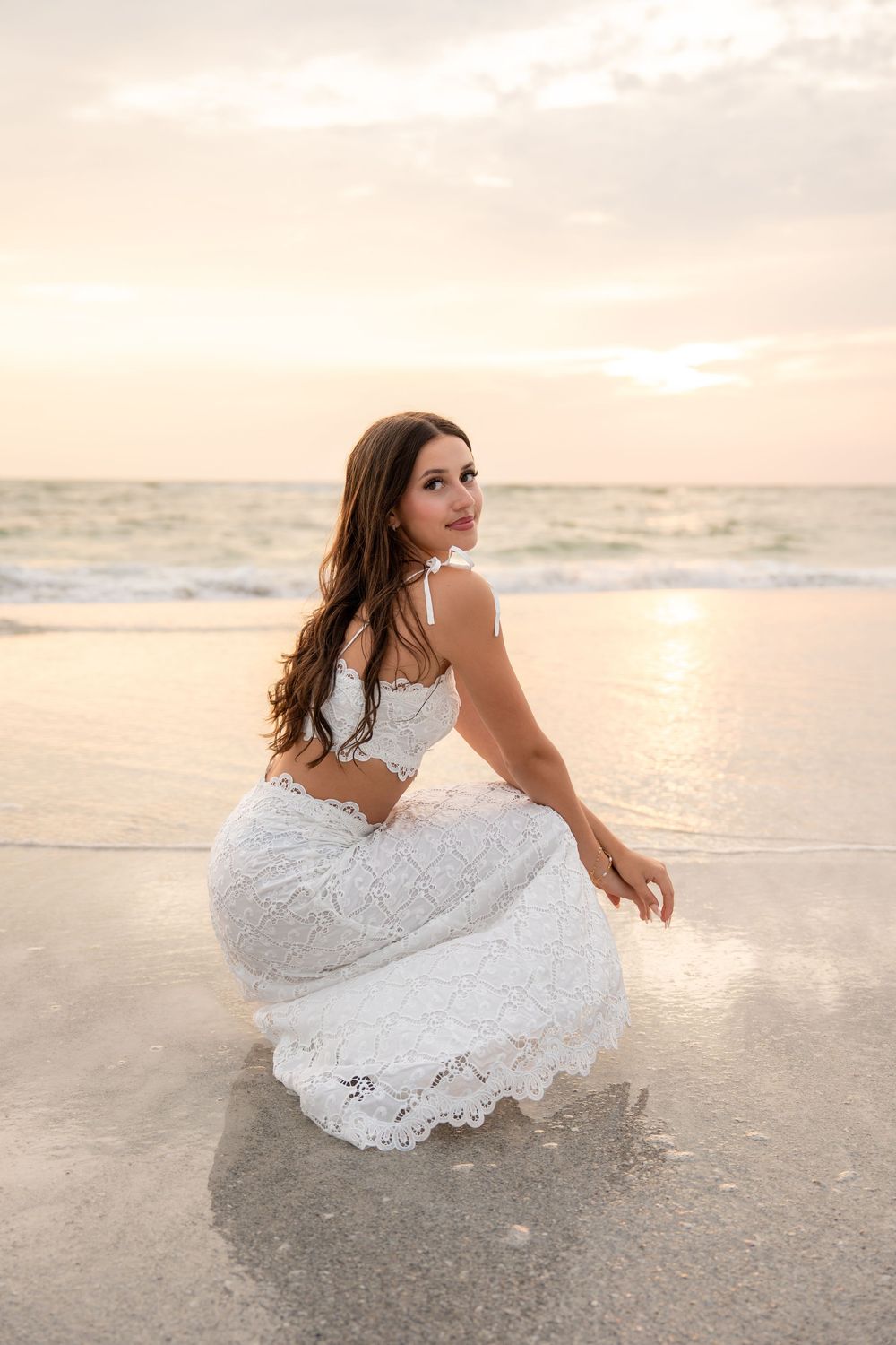 Young woman in white lace dress crouching at the ocean's edge on a sandy beach at sunset.