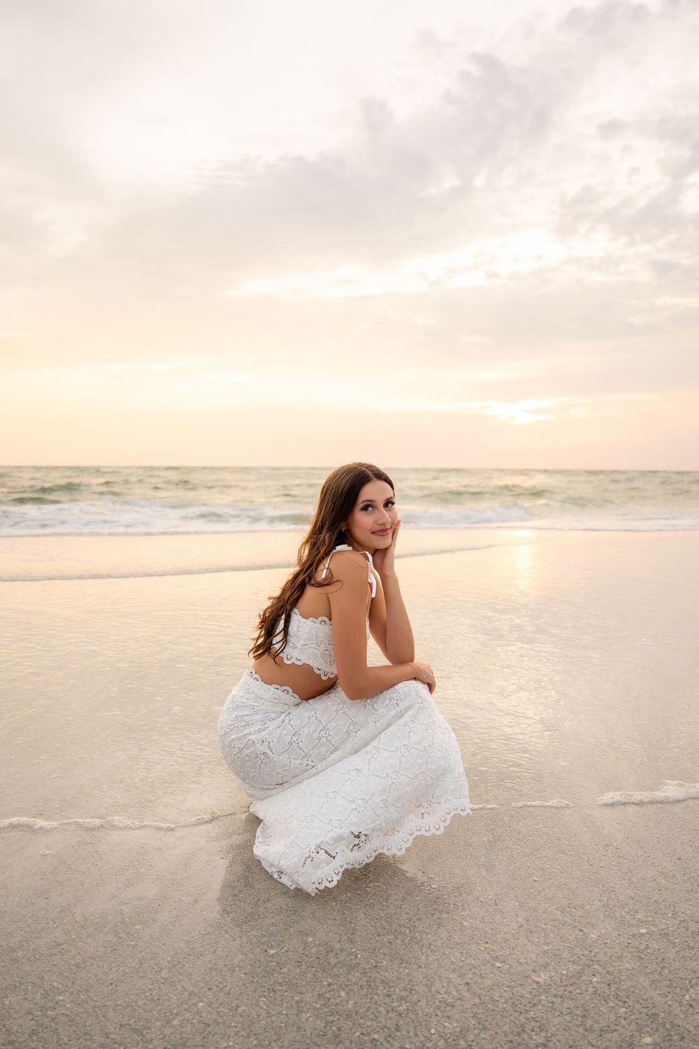 Brunette woman in white lace dress posing on wet sand at the beach during a pastel sunset.