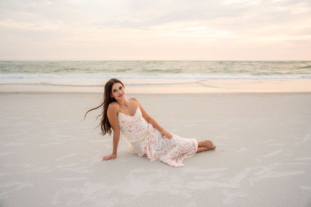 Young woman in white floral dress posing playfully on sandy beach during a golden sunset.