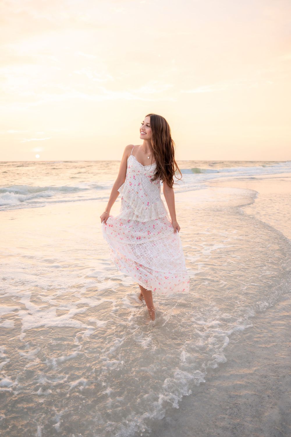 Young woman in floral dress walking barefoot on beach shore at golden sunset.
