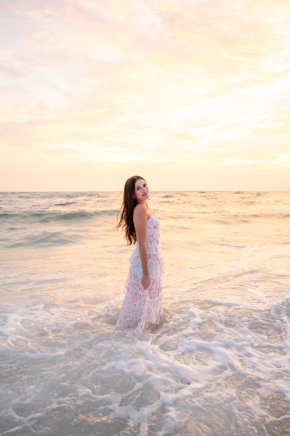 Woman in white floral dress standing in ocean waves during golden sunset.