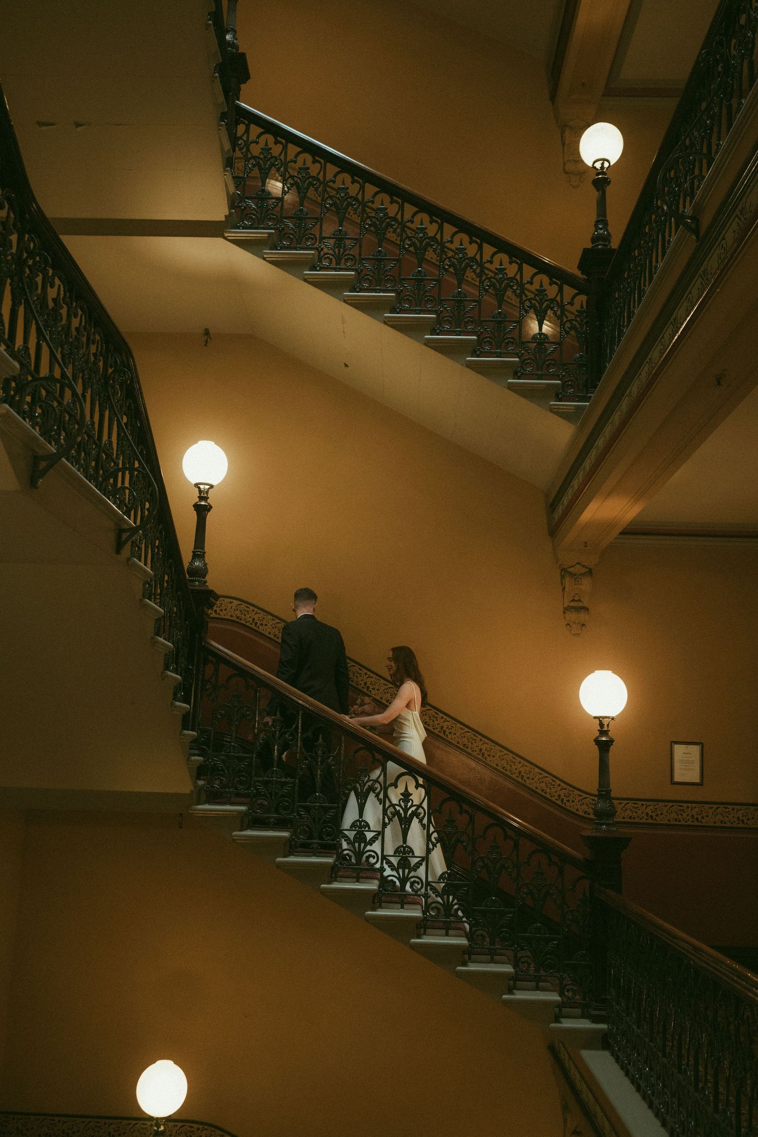 Bride and groom share a romantic moment on an ornate staircase, bride holding pink bouquet in elegant white gown.