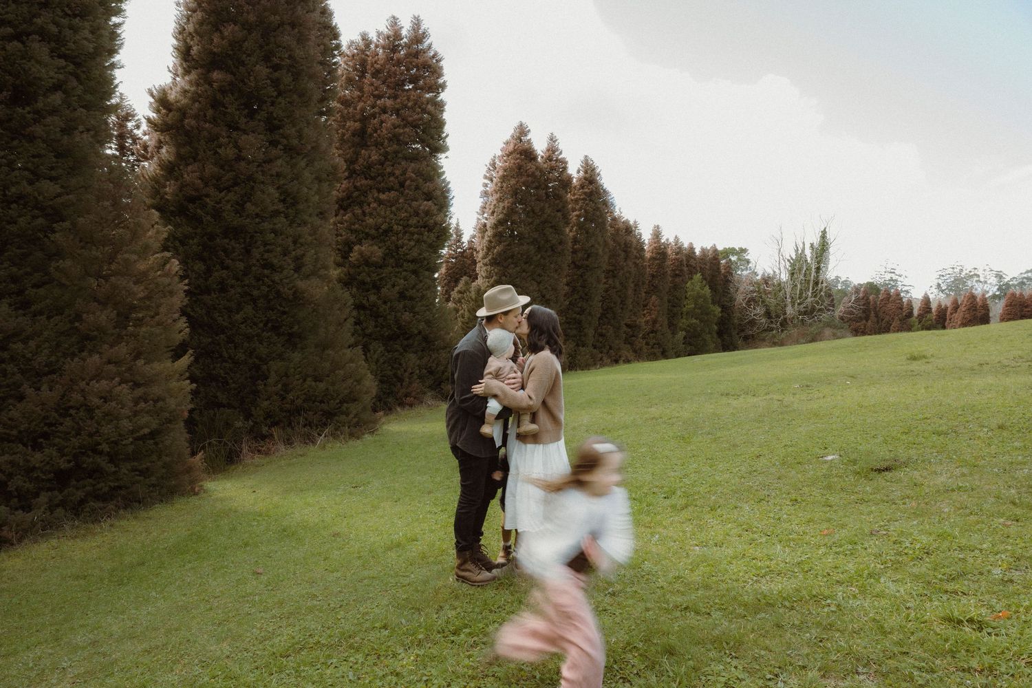 Couple sharing a kiss in a lush green park with tall cypress trees while a child runs in the foreground.
