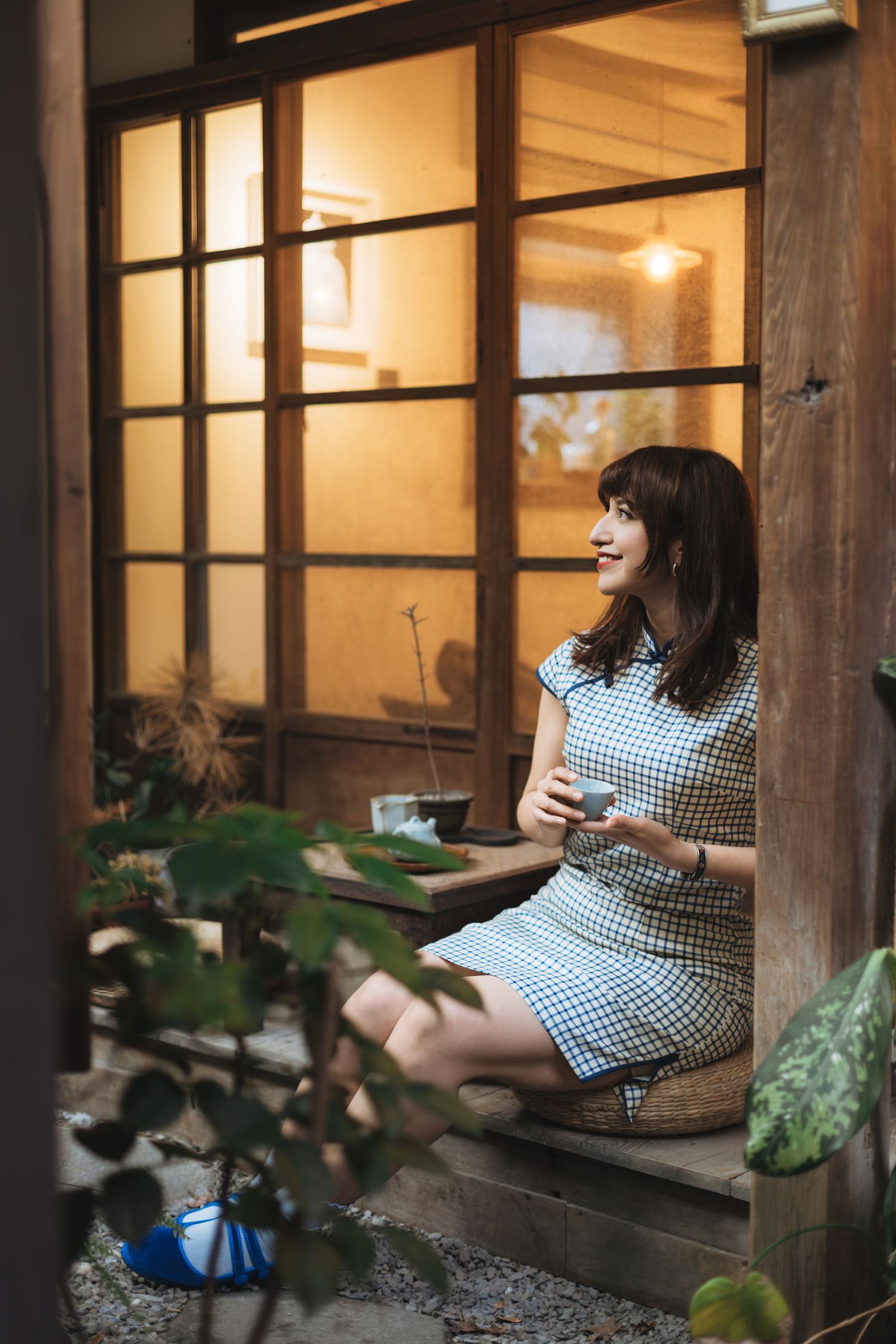 A smiling young woman in a blue gingham dress sits on a wooden step holding a cup outside a warmly lit Japanese cafe.