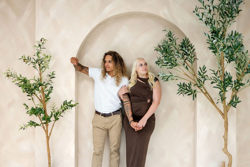 Couple holding hands under arch between olive trees, man in white shirt, woman in brown dress.