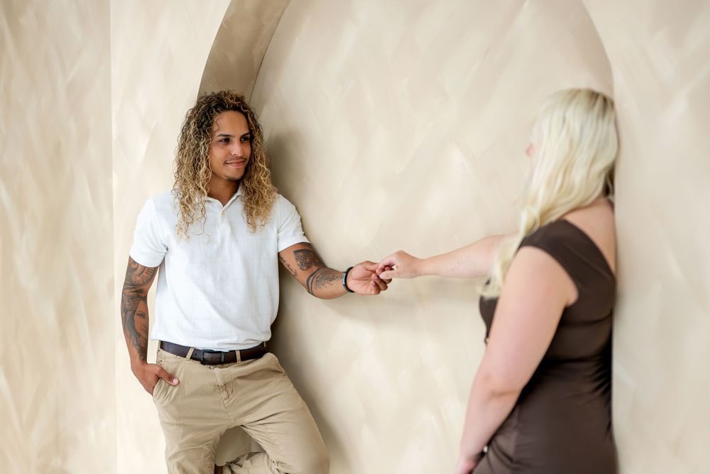 A tattooed man with curly hair holds hands with a blonde woman against a beige arched wall.