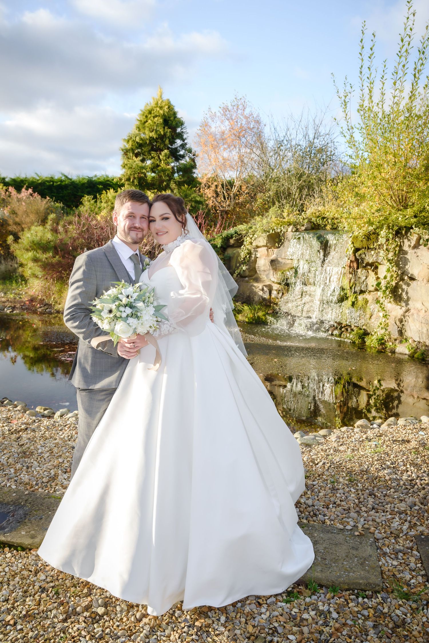 Bride and groom embracing in a lush green meadow on their wedding day, wearing elegant attire in natural countryside setting.