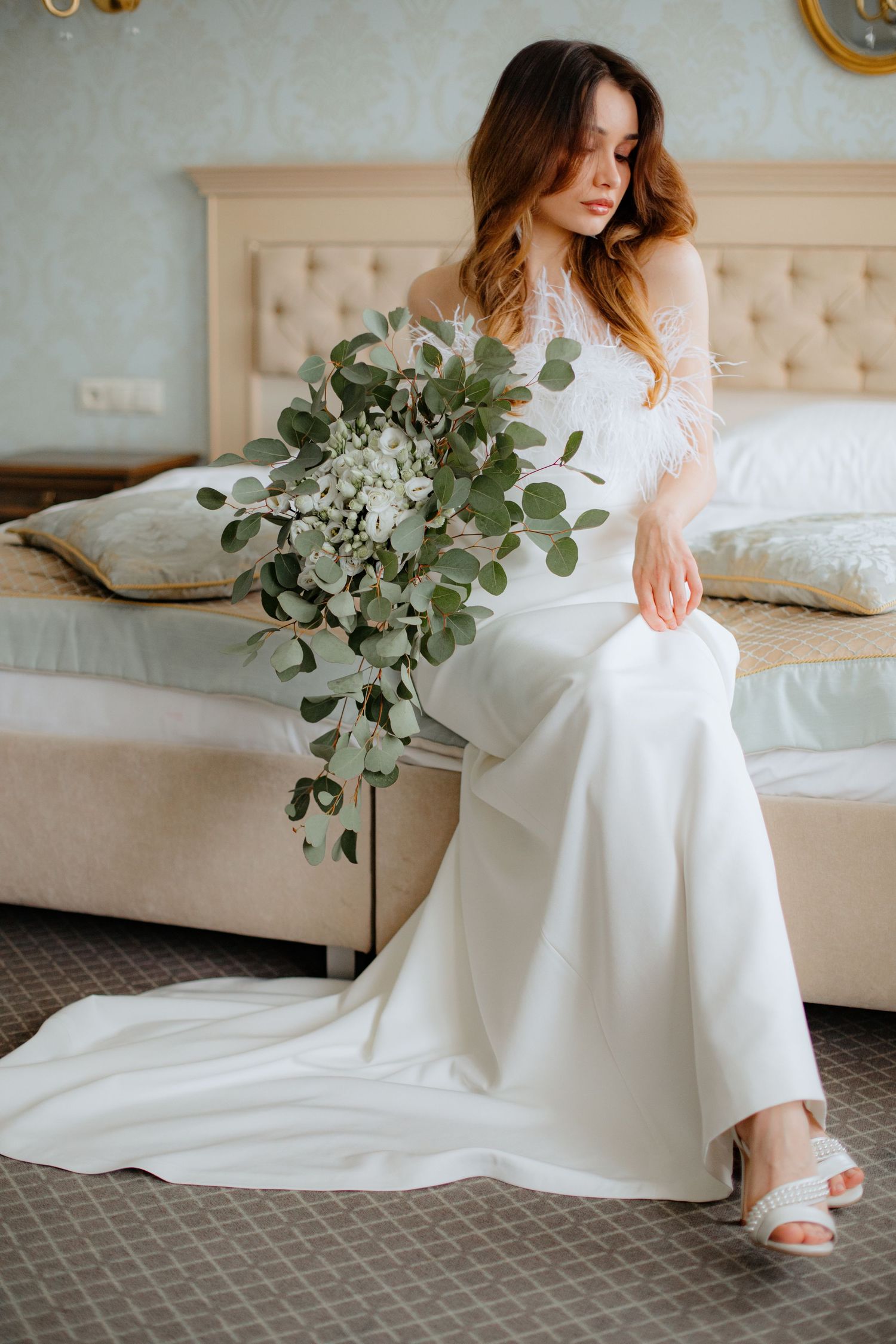 Bride in white lace wedding gown sitting on bed holding cascading eucalyptus bouquet in elegant hotel room.
