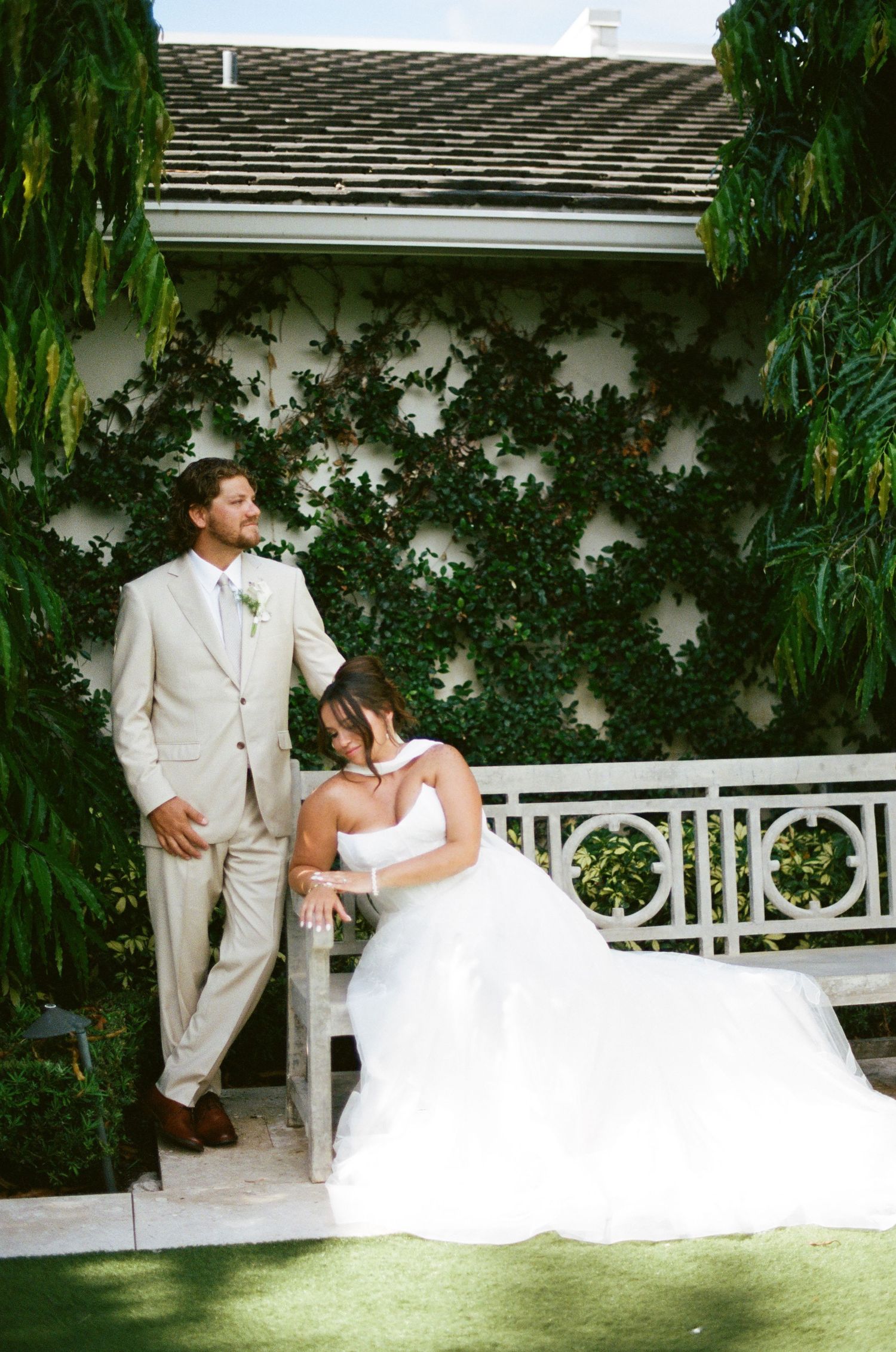 Bride in white gown sits on bench beside groom in tan suit, posing before ivy-covered wall at outdoor wedding.