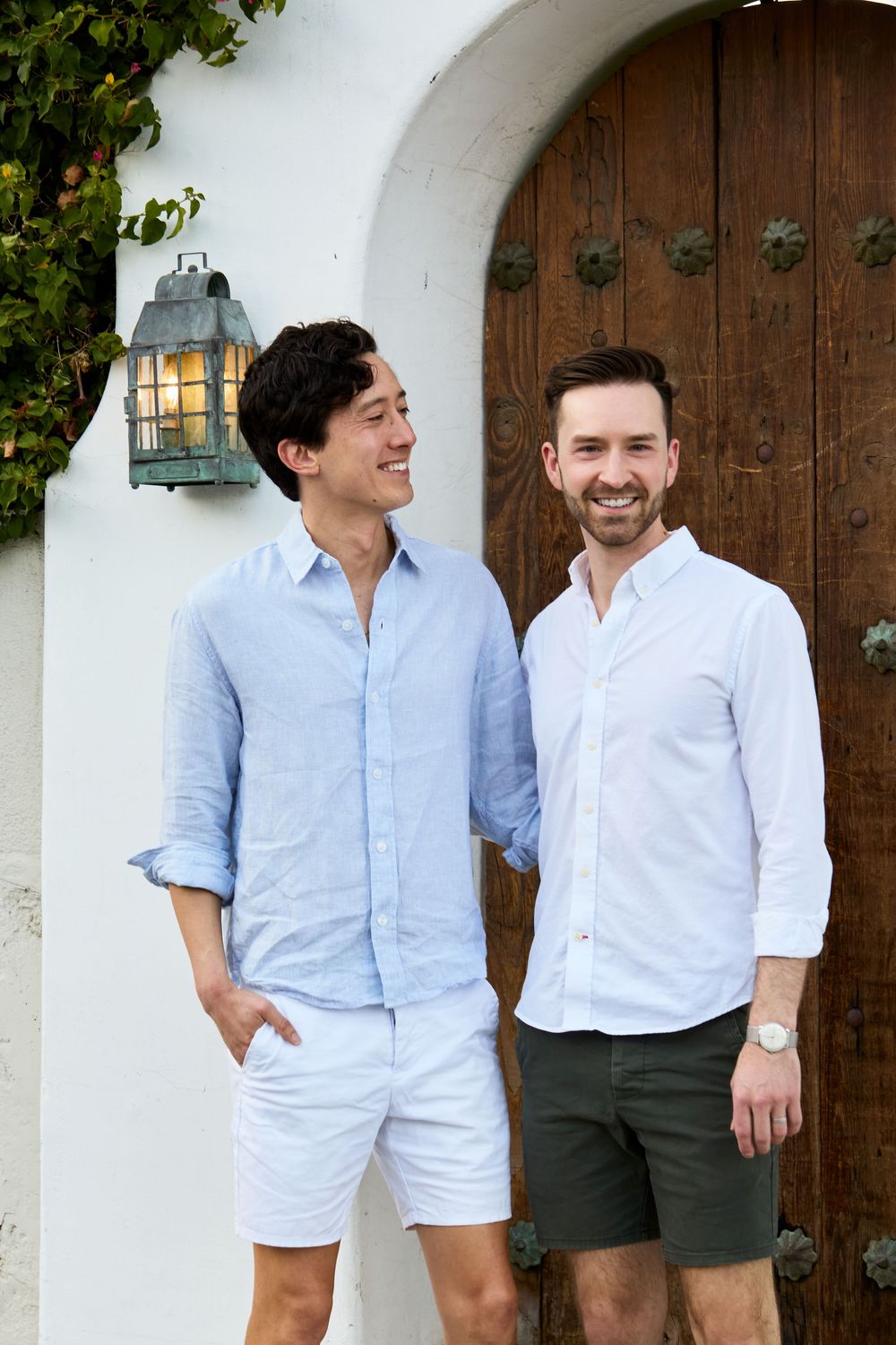 Two smiling men in casual button-up shirts and shorts standing together in front of a rustic wooden door.