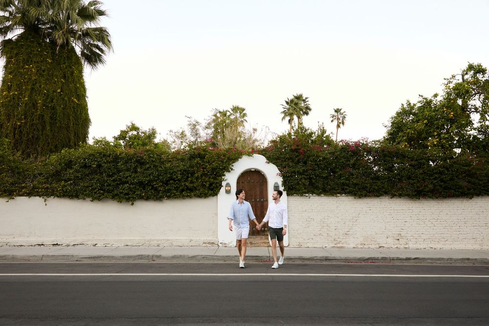 Couple holding hands walking toward an arched white gate surrounded by lush greenery and palm trees on a sunny day.
