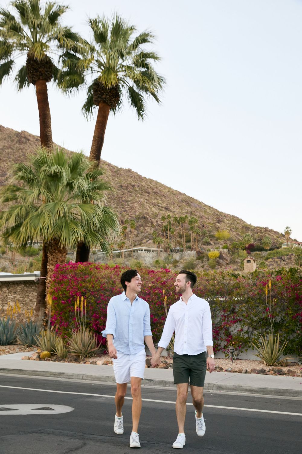 Two men holding hands and walking on a sunny street in Palm Springs with palm trees and colorful flowers.