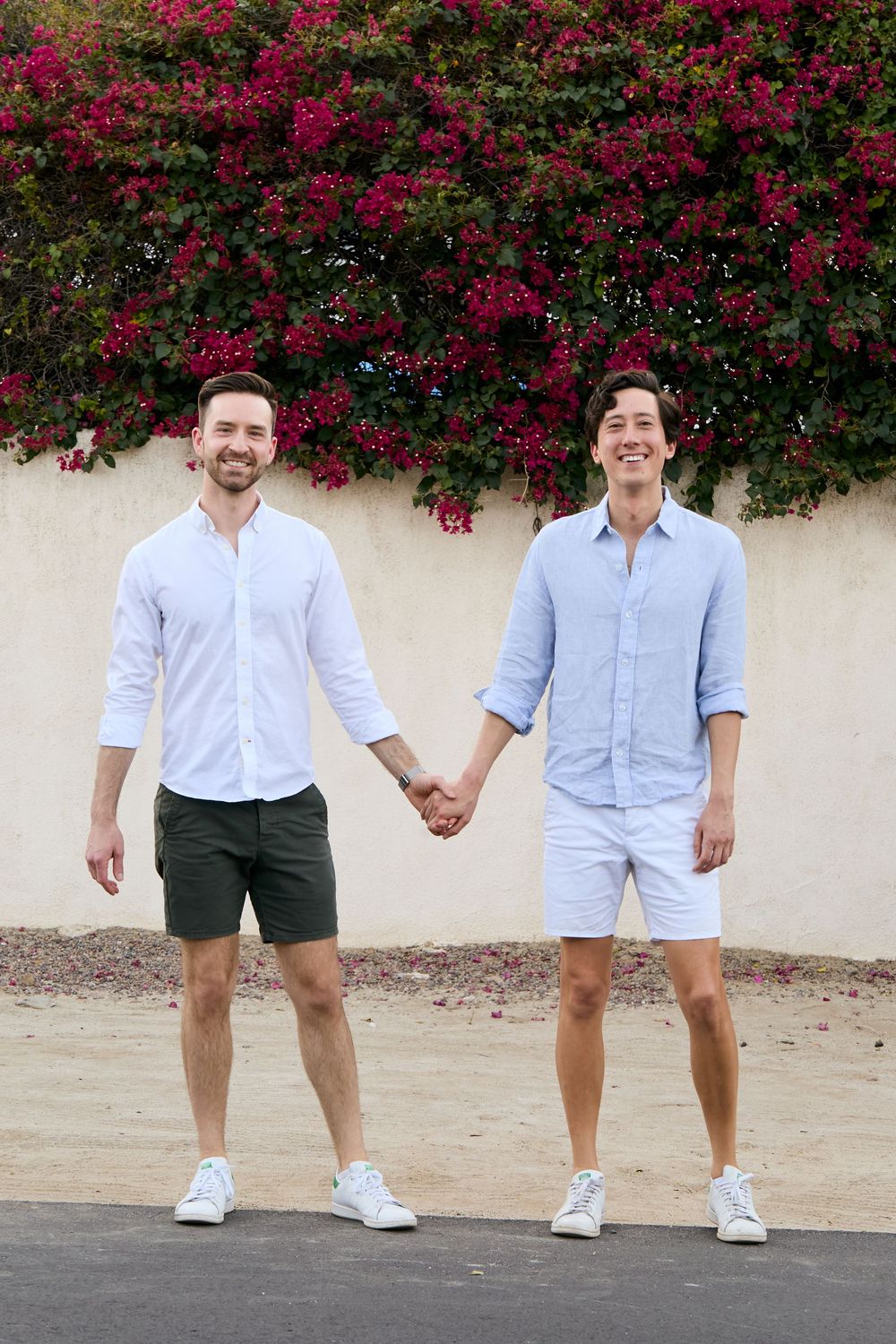 Two men holding hands and smiling outdoors, wearing light blue shirts and shorts, with pink bougainvillea blooms behind them.
