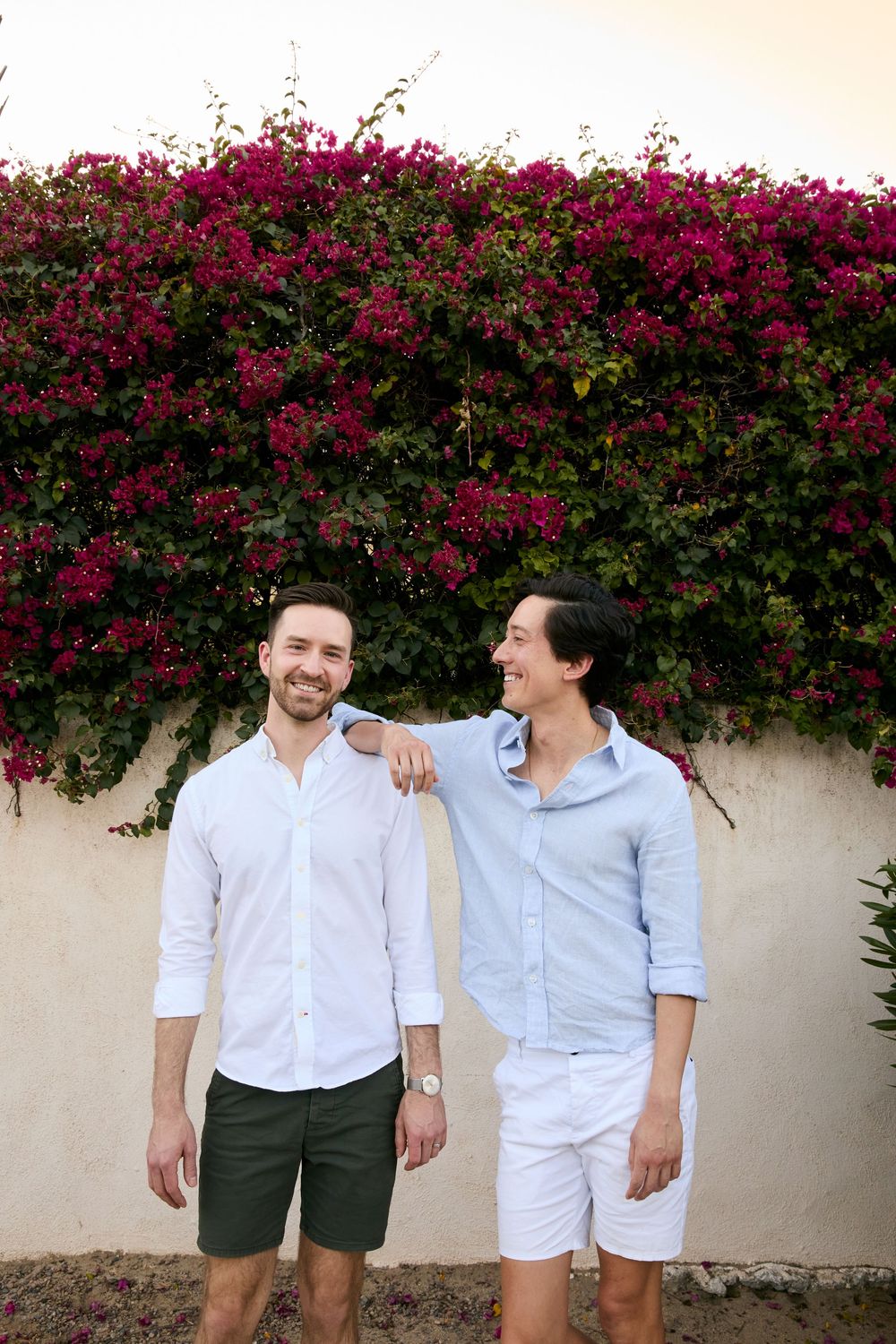 Two smiling men in casual shirts pose together in front of a vibrant bougainvillea wall outdoors.