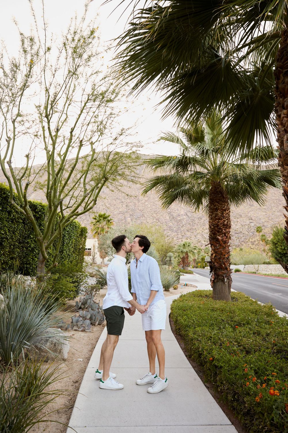 Two men sharing a kiss on a palm-lined sidewalk in Palm Springs with desert mountains in the background.