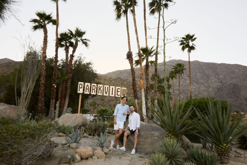 Couple posing by Parkview sign surrounded by palm trees and cacti with mountain backdrop at dusk.