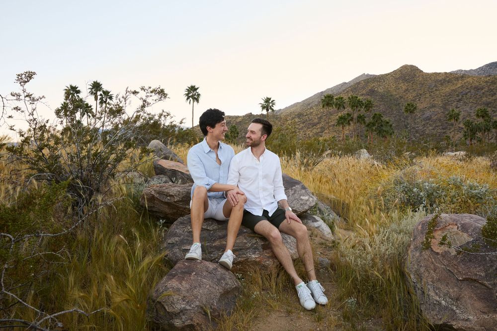 Two men sitting together on rocks in a scenic desert landscape with mountains and palm trees at sunset.