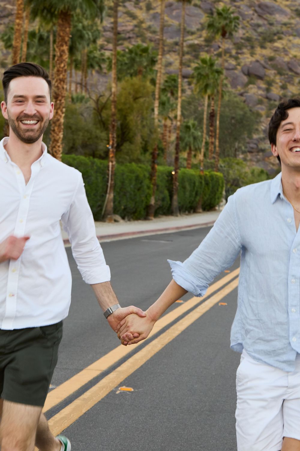 Two smiling men holding hands while walking on a palm tree-lined road in Palm Springs.