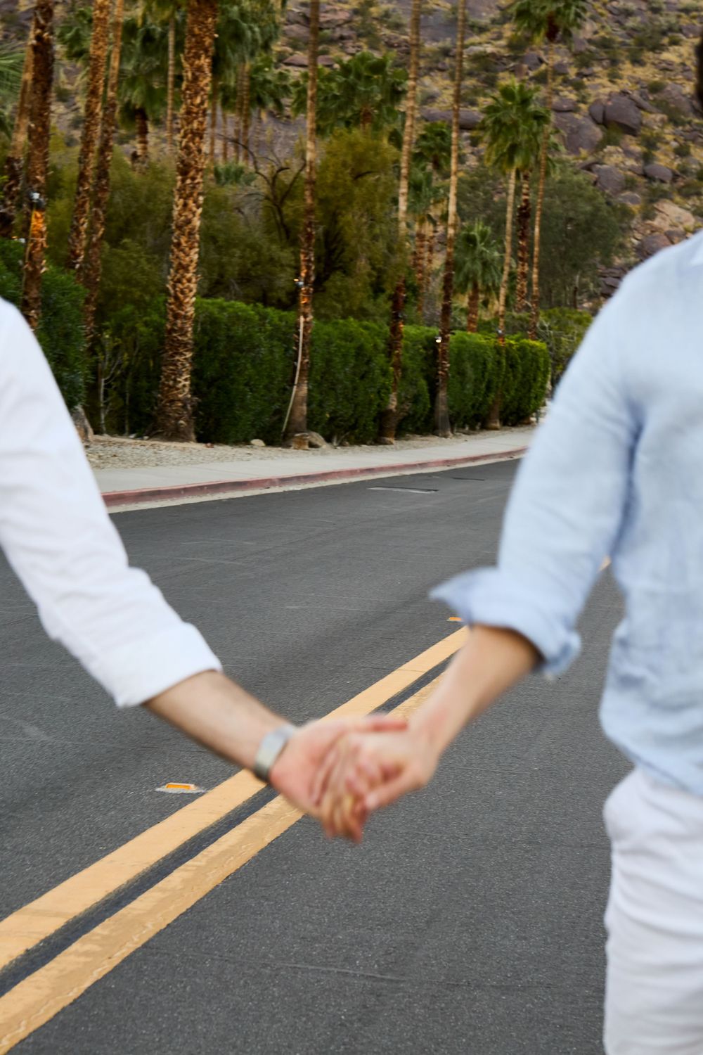 Couple holding hands on a palm tree-lined road in Palm Springs, wearing white and blue shirts.