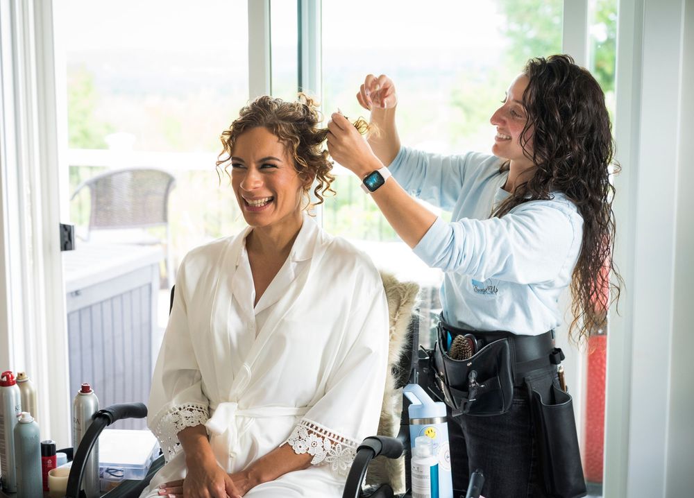 Bride in white robe smiling as hairstylist styles her curly hair on wedding day.