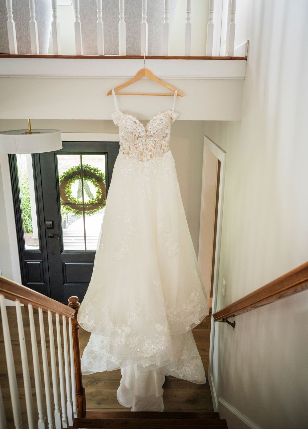 Elegant lace wedding gown hanging on a wooden hanger over a staircase railing near a front door with wreath.