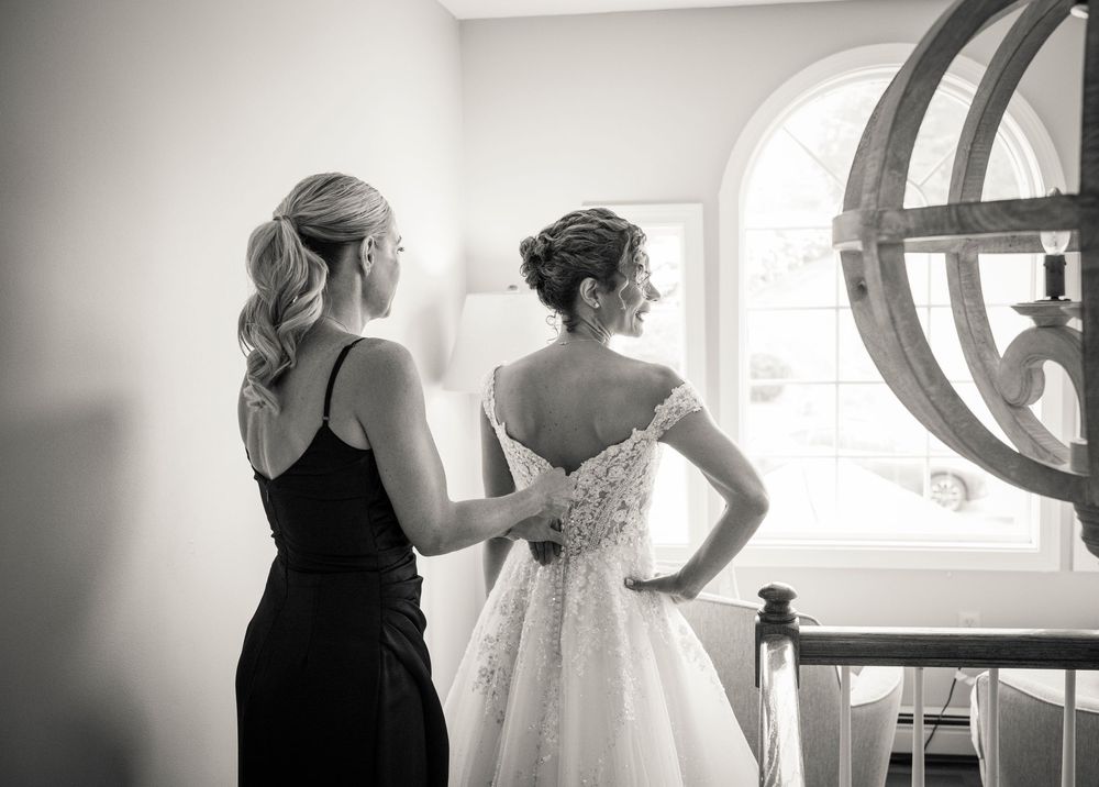 Bridesmaid helping bride with lace wedding dress near bright window in black and white photo.
