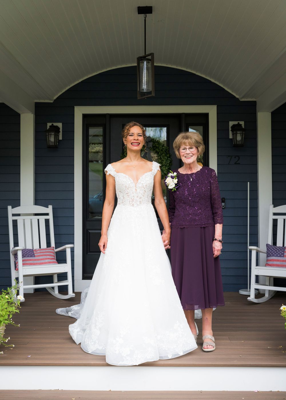 Bride in white lace gown poses with mother in purple dress on blue house porch.