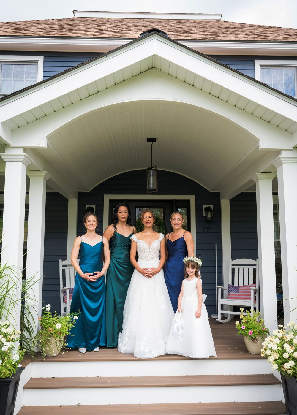 Bride poses with bridesmaids in teal gowns and flower girl on a navy blue house porch.