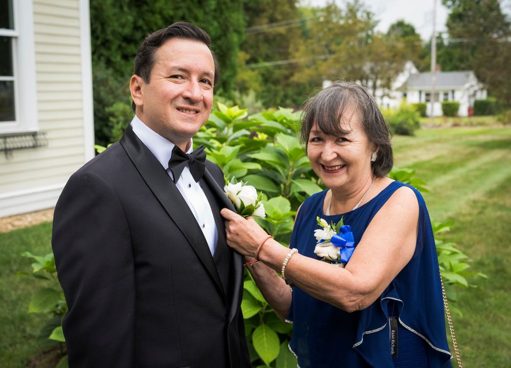 An older woman pins a boutonniere on a man in a tuxedo outdoors at a wedding ceremony.