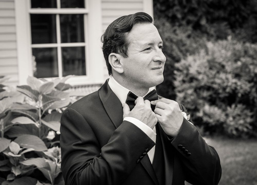 Groom in black tuxedo adjusting bow tie outdoors in black and white wedding photo.