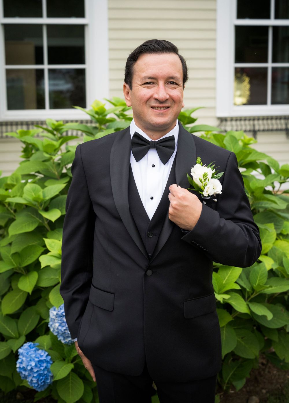 A dapper groom poses in a black tuxedo with bow tie and white boutonniere in front of hydrangea bushes.