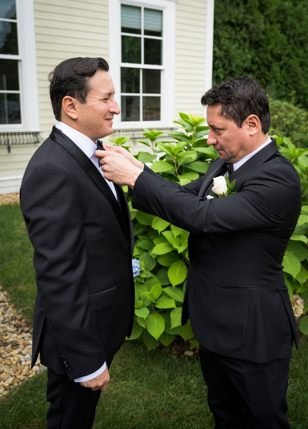 A groomsman adjusts the bow tie of a groom in a black tuxedo outdoors before a wedding ceremony.