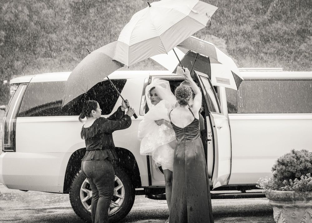 Bride arriving in white limo in the rain, assisted by bridesmaids holding white umbrellas on her wedding day.
