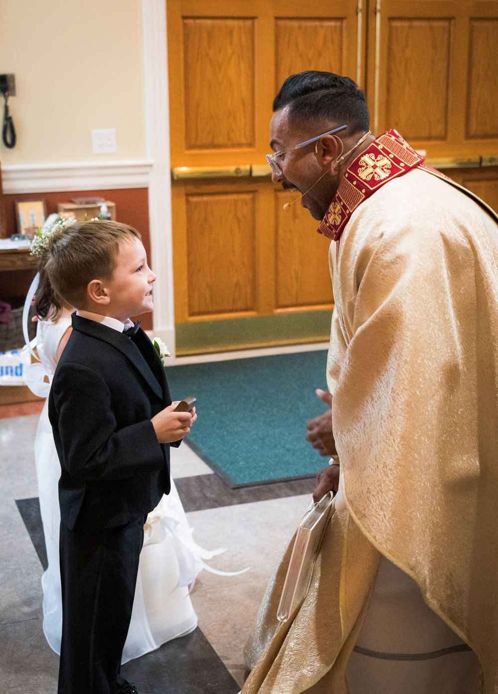 A young ring bearer in a tuxedo conversing with a priest in golden vestments at a church wedding.