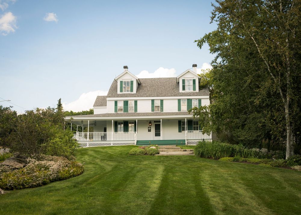 White farmhouse with green shutters and wraparound porch surrounded by lush green lawn and mature trees.