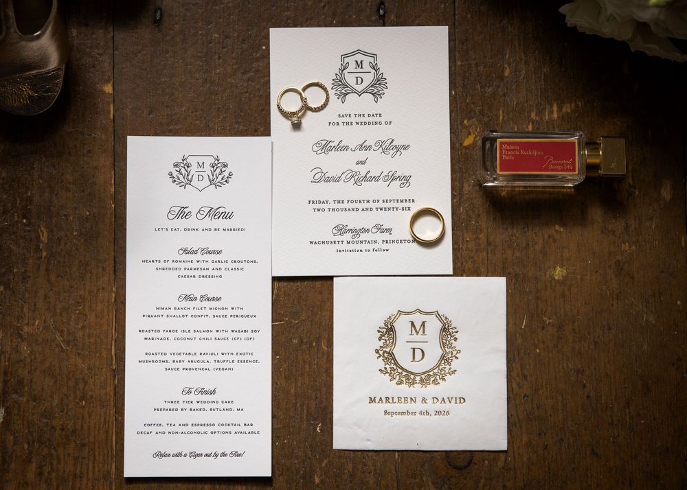 Elegant wedding invitation suite with monogram crest, menu card, and gold rings on wooden surface.