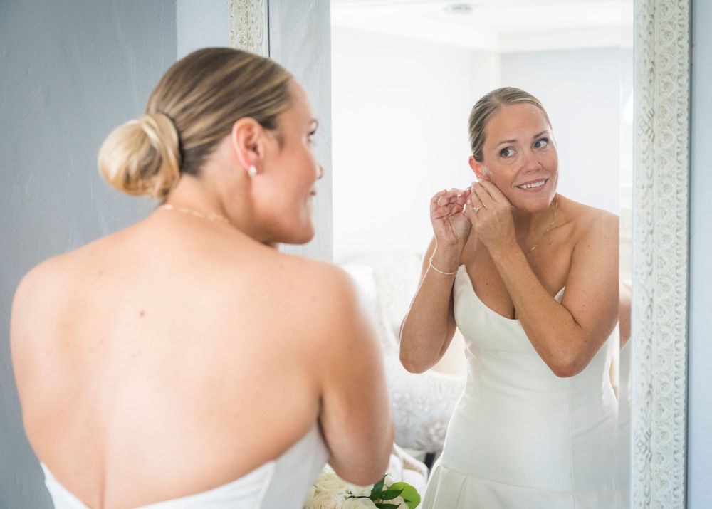 Smiling bride in white towel puts on earrings while looking at her reflection in a mirror.