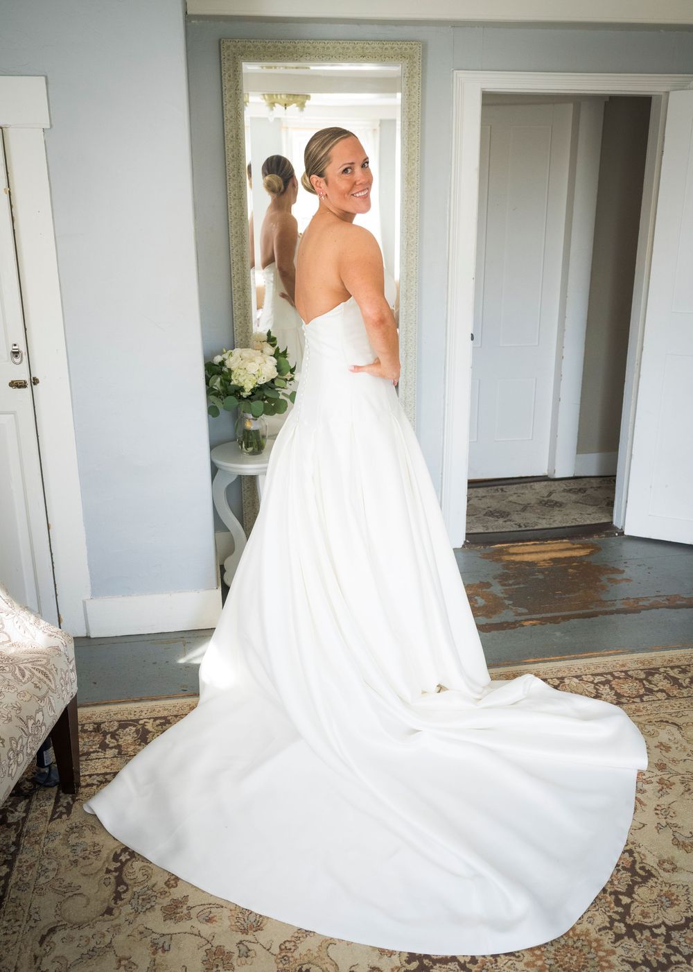 Bride in elegant strapless white ball gown with long train, smiling over shoulder near mirror in soft blue room.