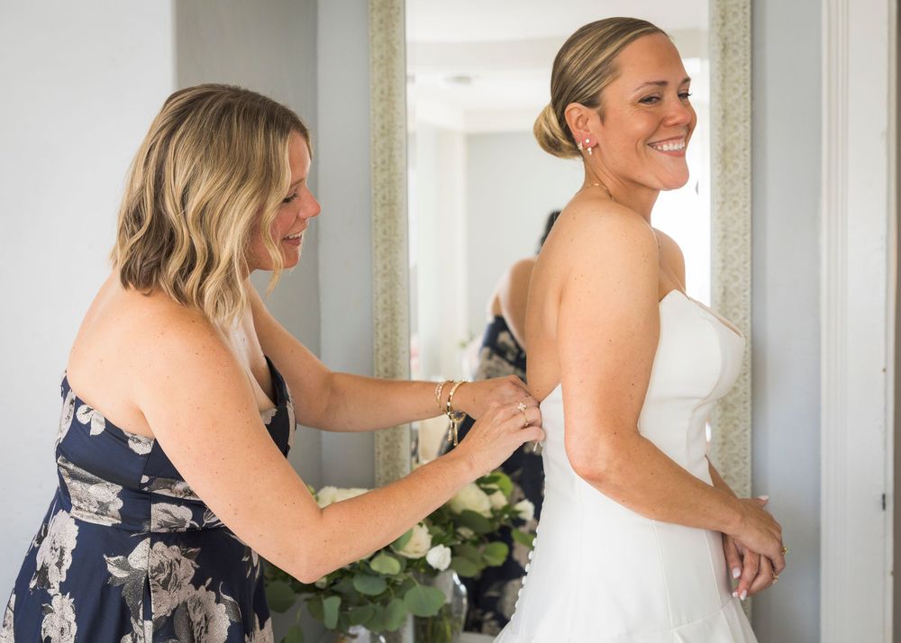 Bridesmaid helping bride zip up her white wedding gown while smiling in front of a mirror.