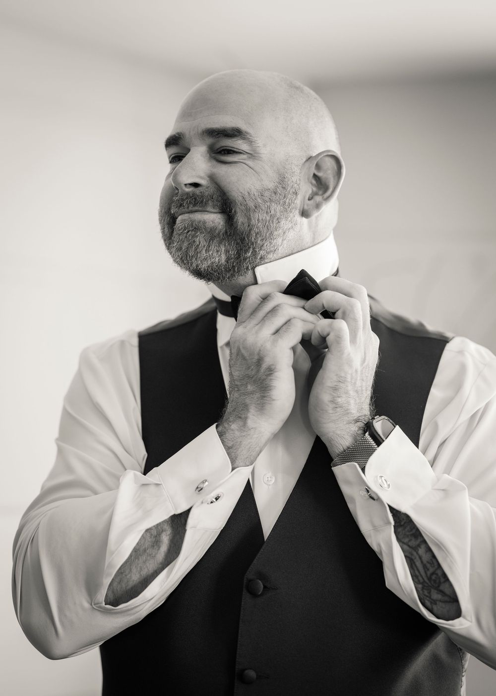 Bald groom with beard adjusting bow tie in black and white wedding portrait.