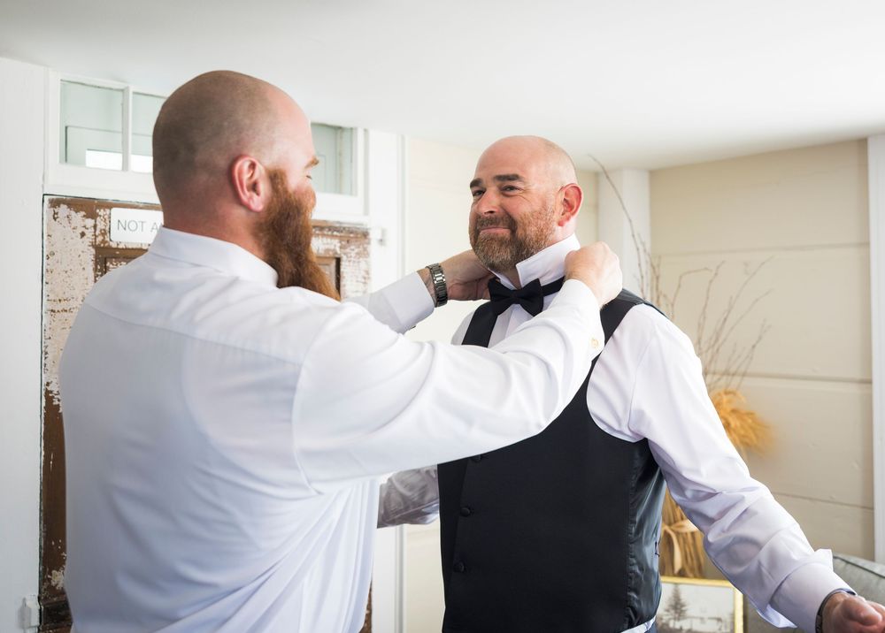 Groomsman helping bald groom adjust bow tie before wedding ceremony in bright preparation room.