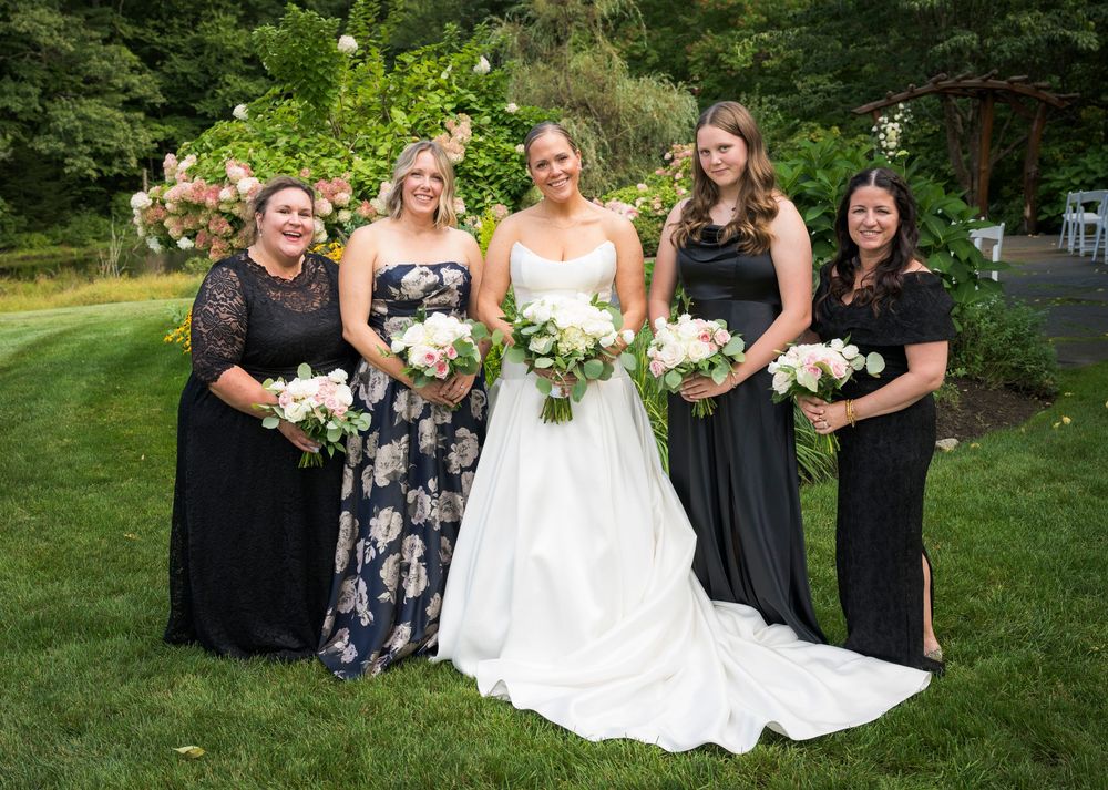 Bride in white gown poses with four bridesmaids in black dresses holding bouquets outdoors at a garden wedding.