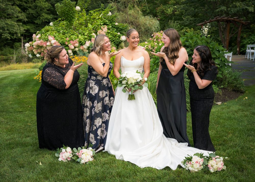 Bride in white gown laughs with four bridesmaids in black dresses on lush green lawn with blooming flowers.
