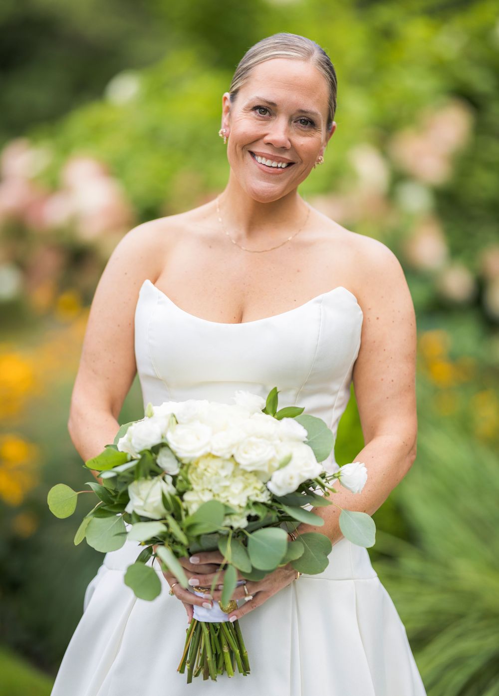 Smiling bride in strapless white gown holding white floral bouquet outdoors.
