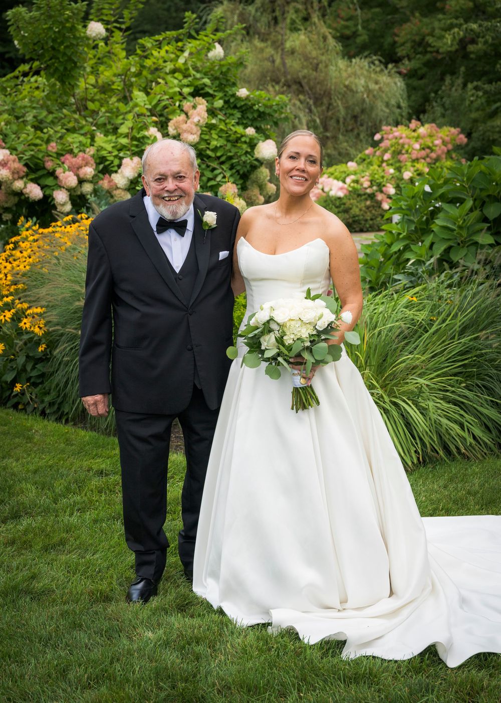 Bride in a strapless white ball gown holding white bouquet poses with older gentleman in tuxedo in garden setting.