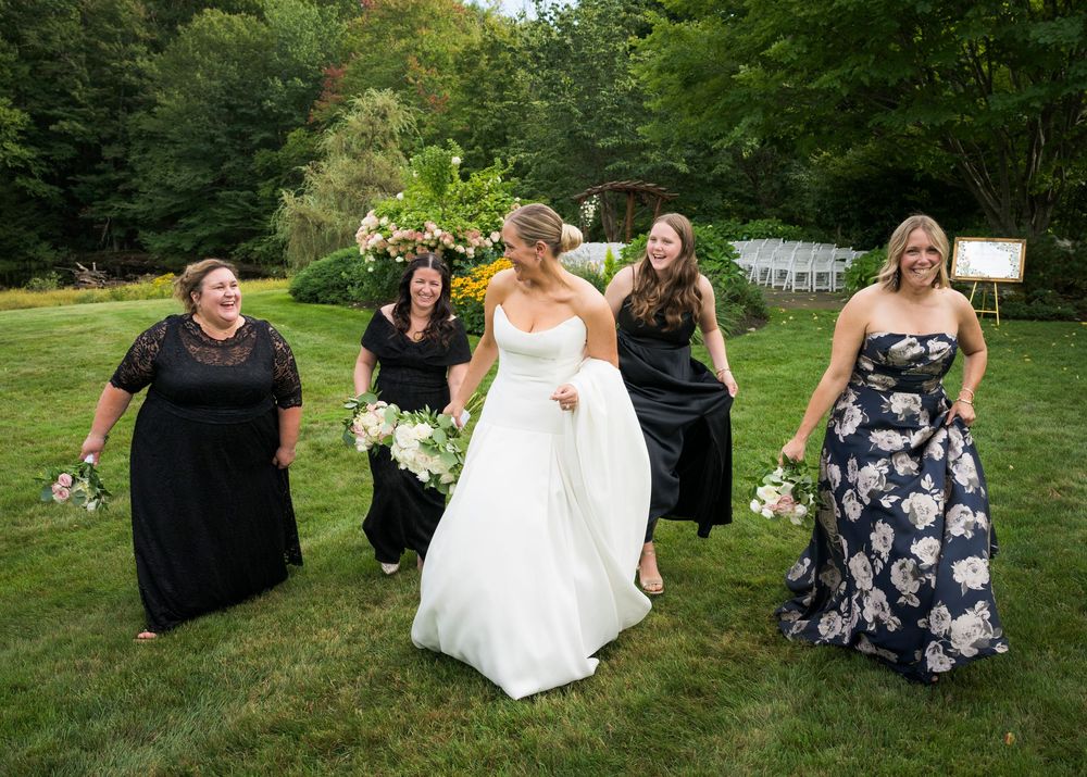 Bride in white gown walks with four bridesmaids in black and floral dresses across lush green lawn.