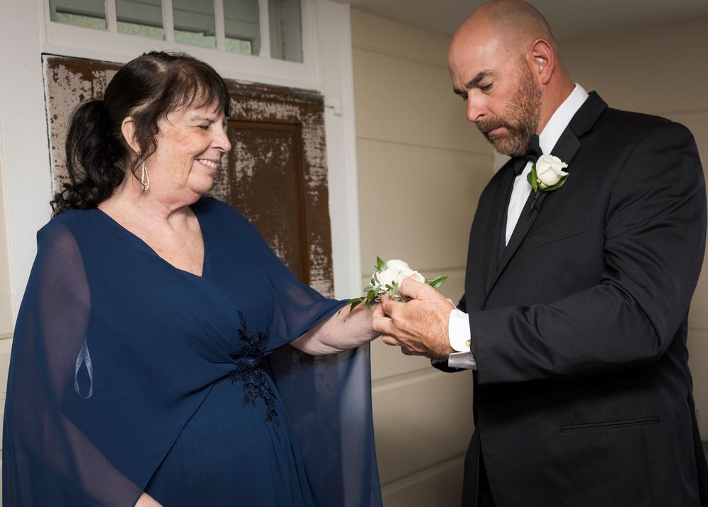 Woman in blue gown pins boutonniere on tuxedo-clad groom before wedding ceremony indoors.