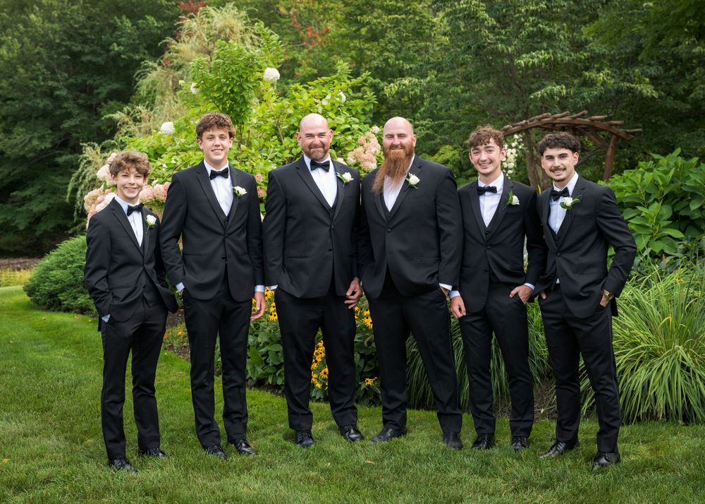 Groom with groomsmen and young men in matching black tuxedos posing outdoors in lush green garden setting.